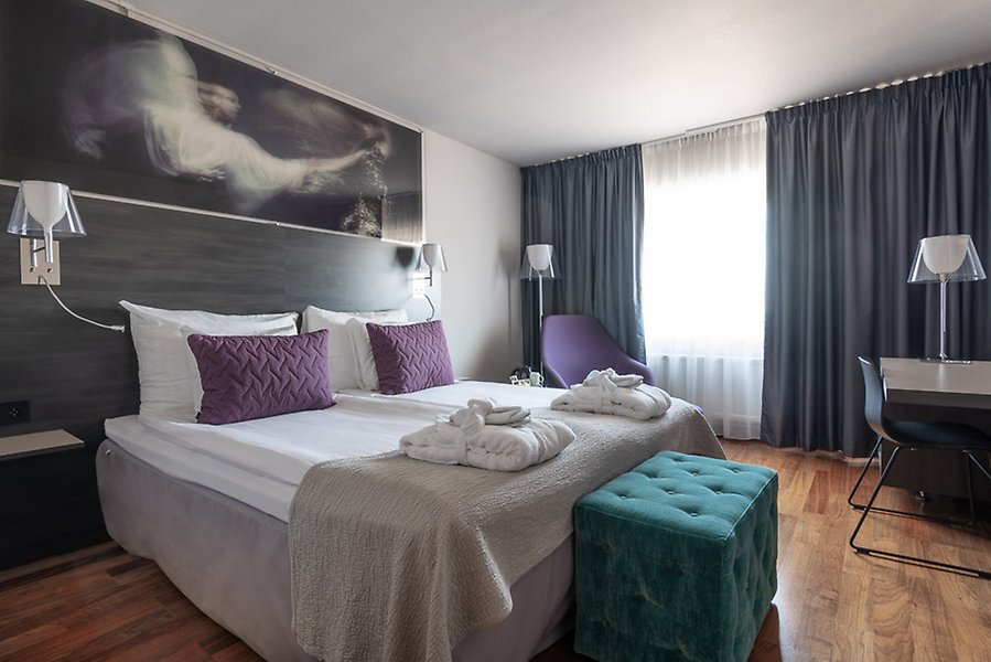 Fotografi av ett hotellrum med en dubbelsäng till vänster i bild och ett skrivbord till höger i bild.