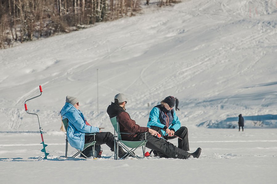 Fotografi av tre personer som sitter i campingstolar på isen och pimplar. Isen är täckt av snö. Personerna har på sig täckbyxor, jackor och mössor. Bakom dem står en isborr.
