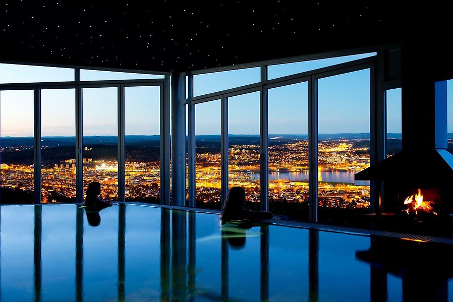 Foto av en inomhuspool med fönster bakom så att man ser ut över Sundsvalls stad. I poolen är det två personer som kolar ut på utsikten bakom poolen.  